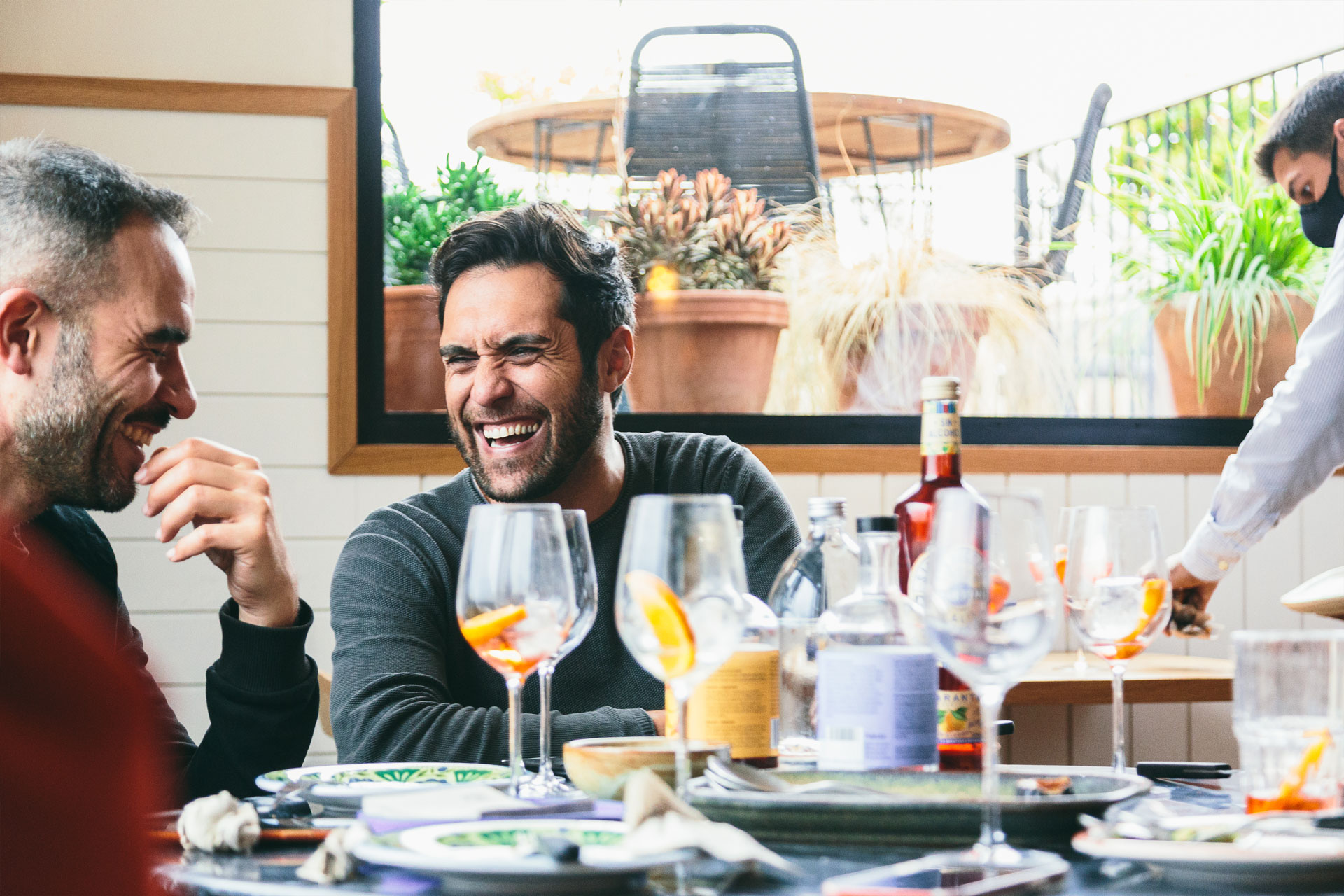 dos hombres riendo en primer plano mientras beben copas de martini rojo con rodajas de naranja y en el fondo se ve el camarero
